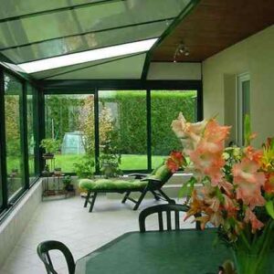 intérieur véranda sur mesure verte avec soubassements, une table avec des fleurs dessus en premier plan et un transat au fond