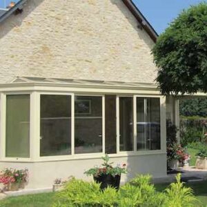 véranda en kit sur mesure de couleur beige avec soubassement avec toiture à chéneaux périmétriques adossée à une façade et arbre à droite